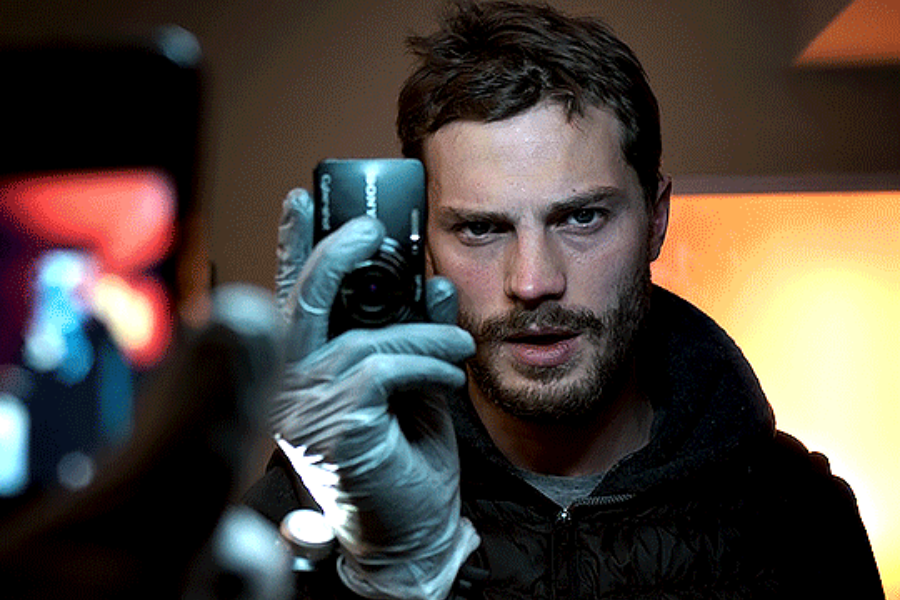 A bearded man wearing gloves holds a digital camera up to his face, capturing a reflection in a dimly lit room.