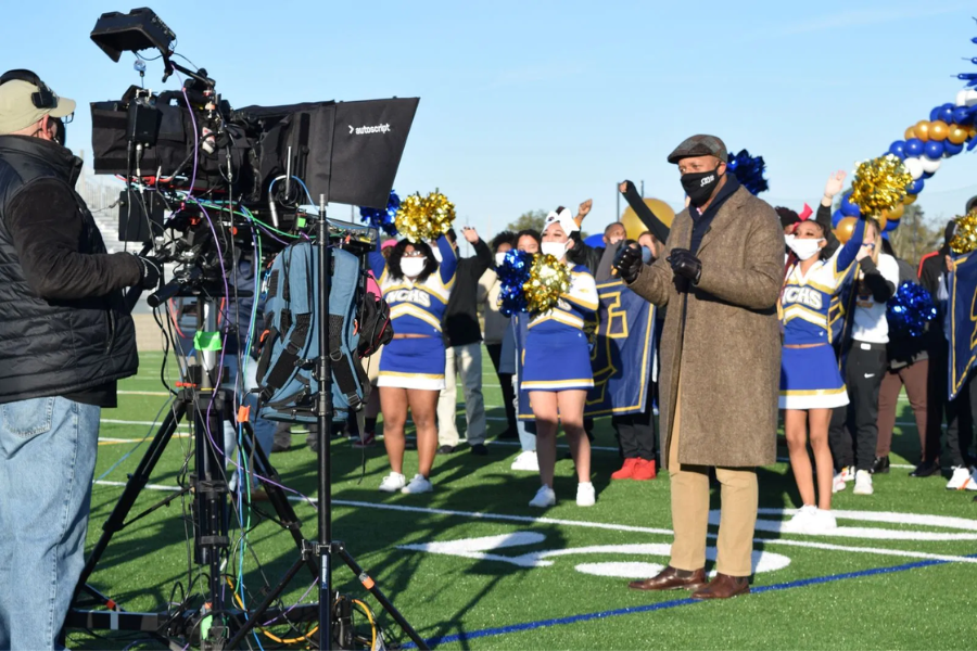 Television crew films a man in a long brown coat speaking on a football field while cheerleaders in blue and gold uniforms cheer behind him.