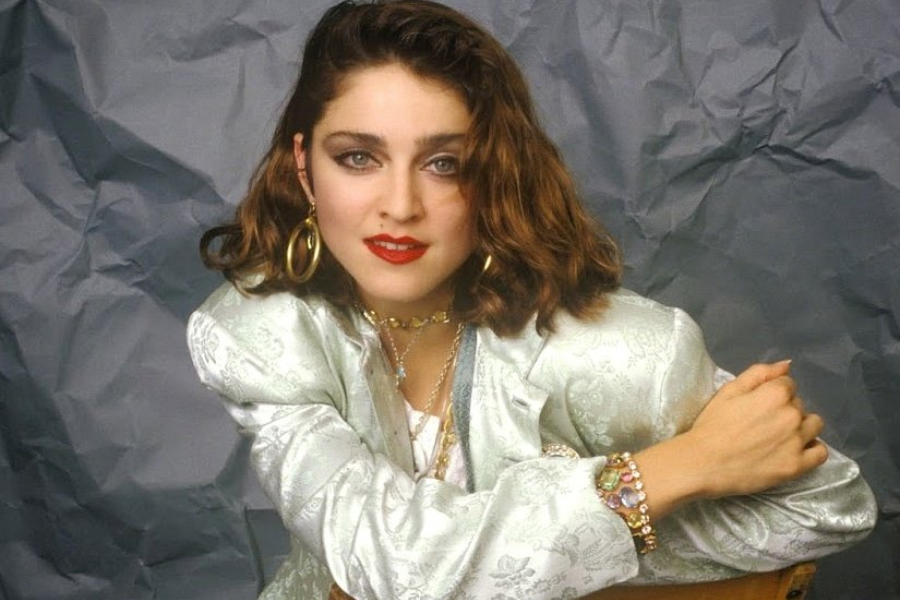 Woman with shoulder-length brown hair wearing a pale brocade jacket and bold jewelry leans forward with a confident expression.