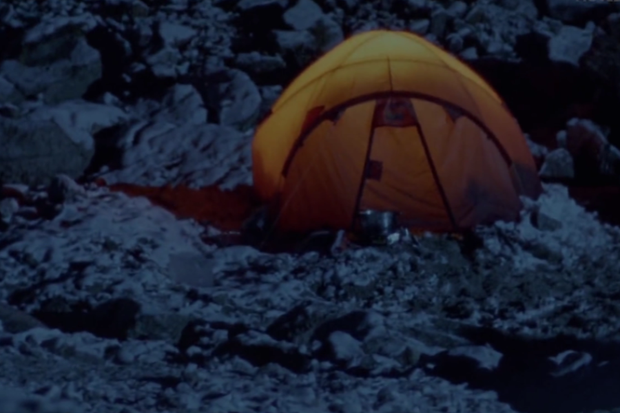 A glowing orange tent stands alone on a snowy, rocky mountainside at night, casting warm light against the cold terrain.