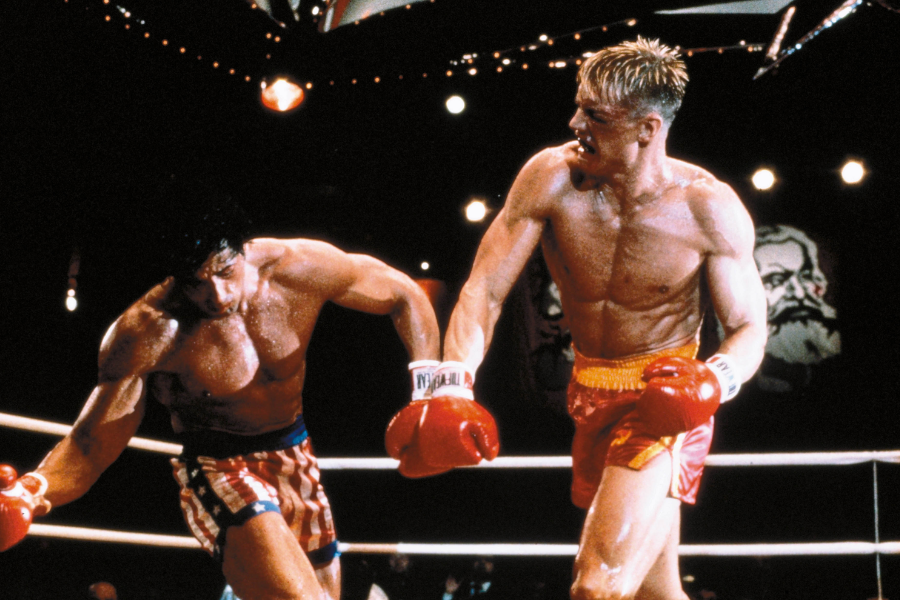 A dramatic boxing match scene with a muscular man in red trunks punching another fighter wearing stars-and-stripes shorts.