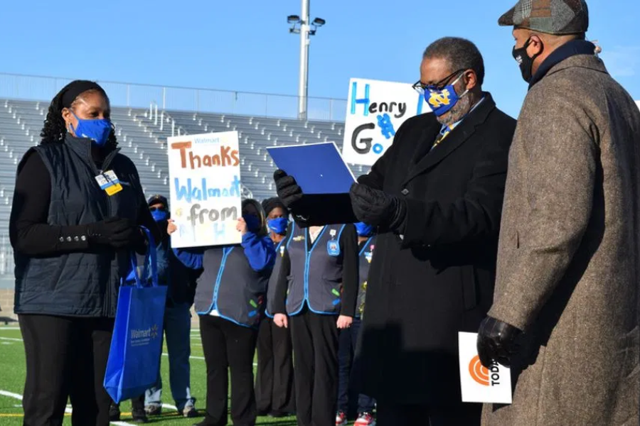 Man in a black coat reads from a document while another man and a Walmart employee stand nearby, with coworkers holding thank-you signs in the background.