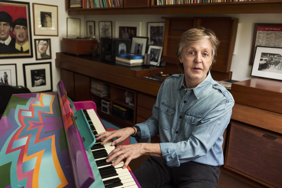 Man in a denim shirt plays a colorful, hand-painted piano in a cozy room decorated with framed photos and records.
