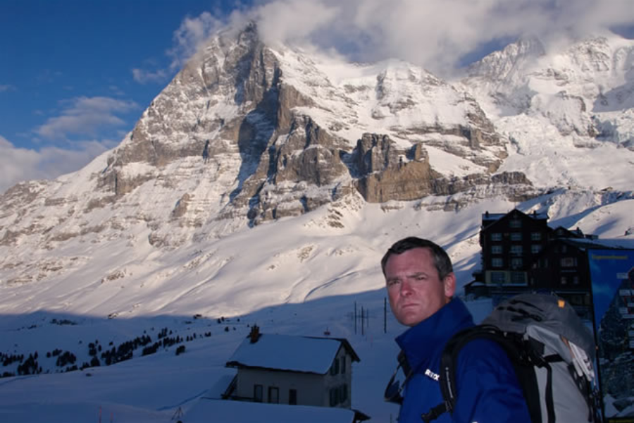 A man in a blue jacket with a backpack stands before the towering snow-covered Eiger mountain in Switzerland under a partly cloudy sky.