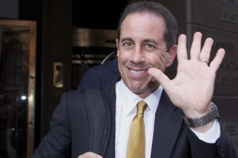 Man in a suit and yellow tie smiles and waves at the camera while standing outside a building.