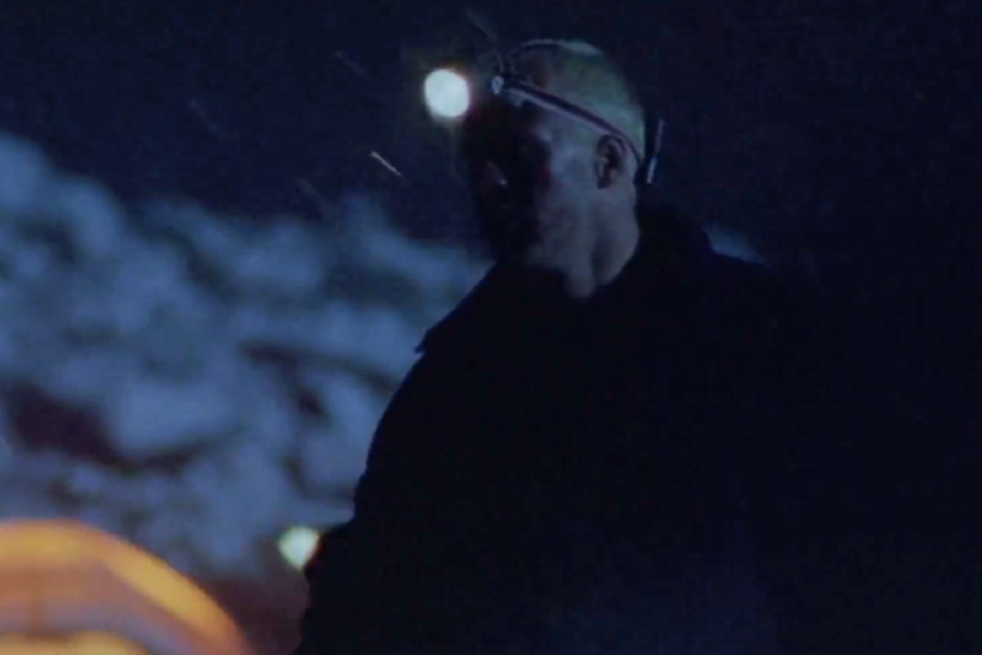 A man wearing a headlamp stands outside at night in snowy conditions, illuminated by dim camp lighting in the background.