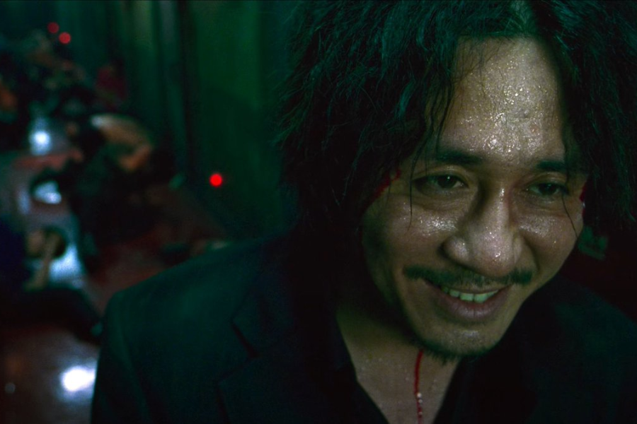 A sweaty man with blood on his face smiles eerily in a dim hallway, with unconscious bodies in the background.
