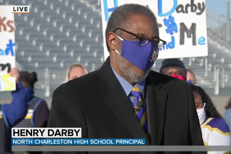 North Charleston High School principal Henry Darby wears a blue and yellow mask while speaking outdoors during a live broadcast.