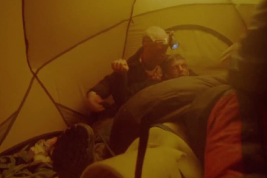 Inside a warmly lit tent, a man supports another who appears weak or injured while lying down, surrounded by gear and sleeping bags.