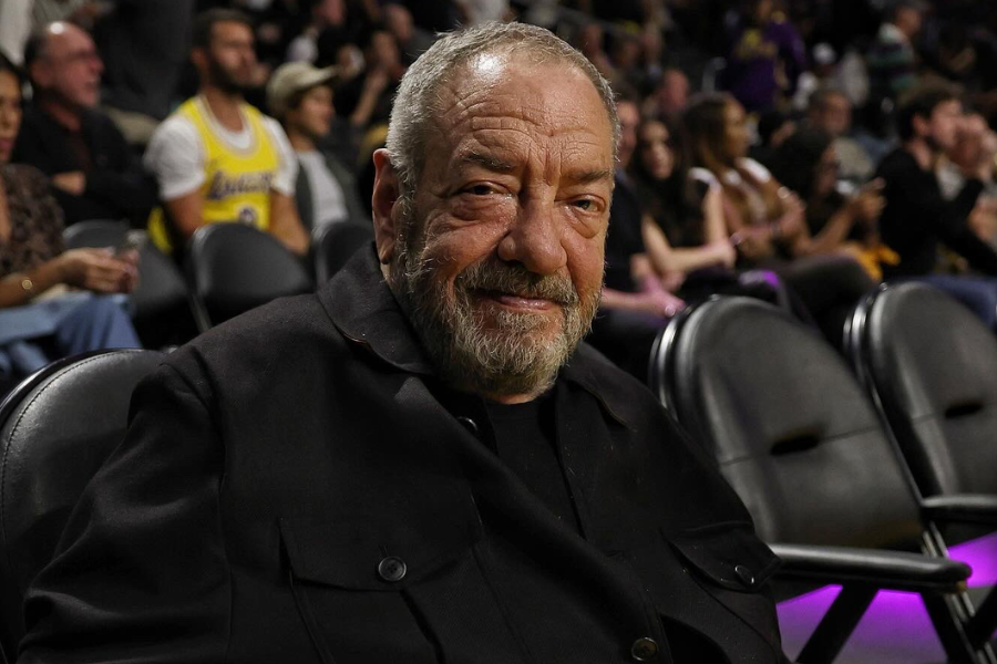 Older man with a beard smiles while seated courtside at a basketball game, with fans in the background.