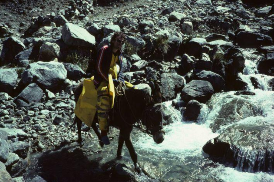 A climber rides a mule across a rocky, shallow stream, surrounded by boulders and mountain terrain, dressed in yellow outerwear.