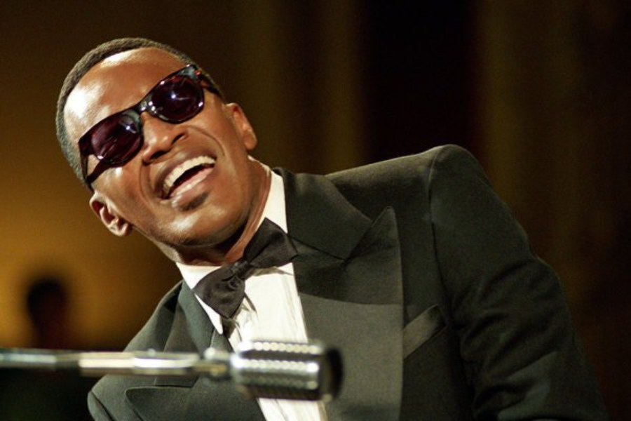 A man in a tuxedo and sunglasses plays piano while smiling joyfully under warm stage lights.