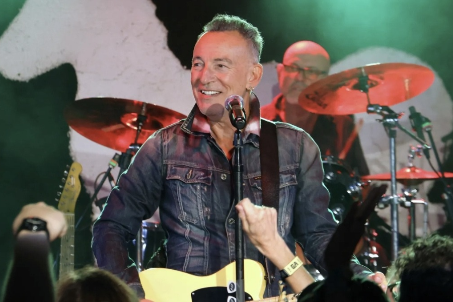 Smiling man in a denim jacket performs on stage with a guitar, as a drummer plays behind him.