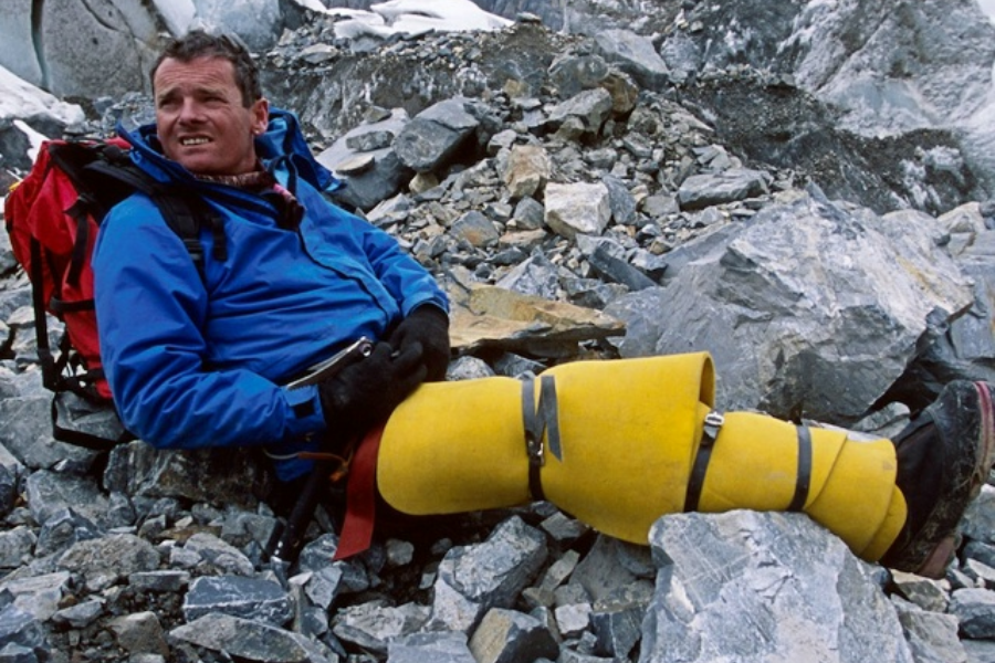 A climber in a blue jacket with yellow leg braces sits on rocky terrain, resting with a red backpack after a strenuous descent.