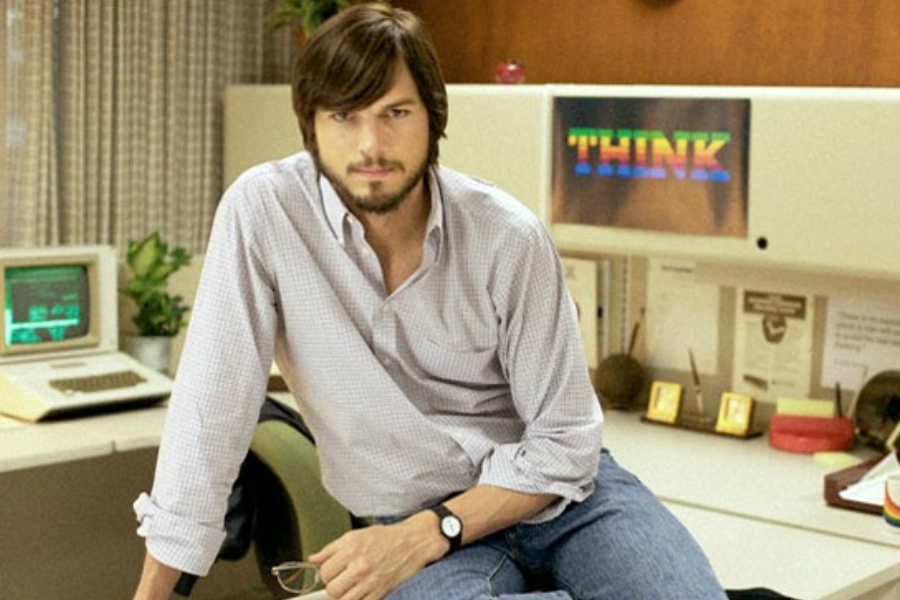 A man with long hair and a beard sits casually on a desk in a retro office space with an old computer behind him.