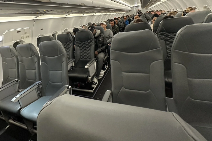 Interior of a commercial airplane showing rows of grey seats and passengers seated toward the back.