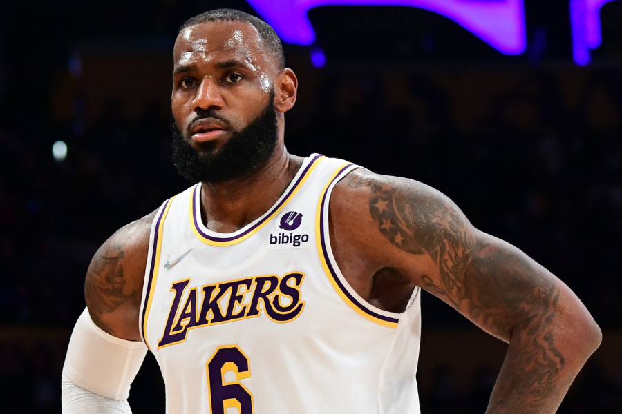 Man in a white Lakers jersey with number 6 stands focused on the basketball court with purple lights in the background.
