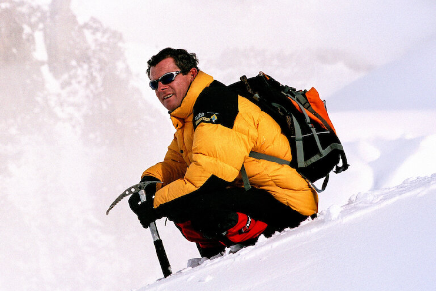 A smiling climber in a yellow down jacket and sunglasses kneels in deep snow with an ice axe, looking out over a white, snowy expanse.