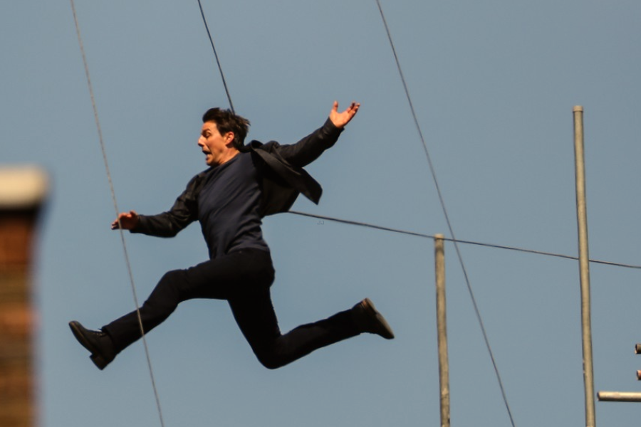 A man in black mid-air during a rooftop jump stunt, suspended by wires against a clear sky.