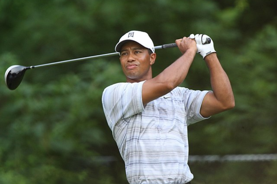 Golfer in a striped shirt and white cap follows through on a swing with a focused expression against a wooded background.