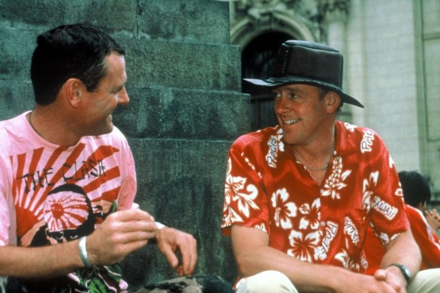 Two men in colorful shirts sit on stone steps laughing together, one wearing a hat and Hawaiian shirt, the other a band shirt.