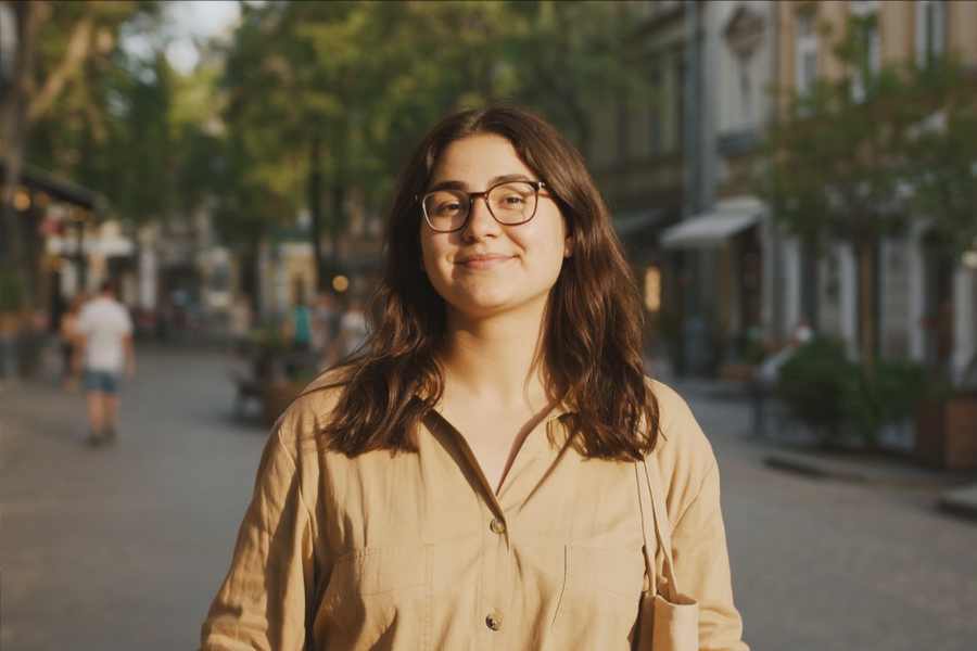 A young woman stands on a sunlit street, smiling gently at the camera with a relaxed expression. She wears a light button-up shirt and carries a tote bag.