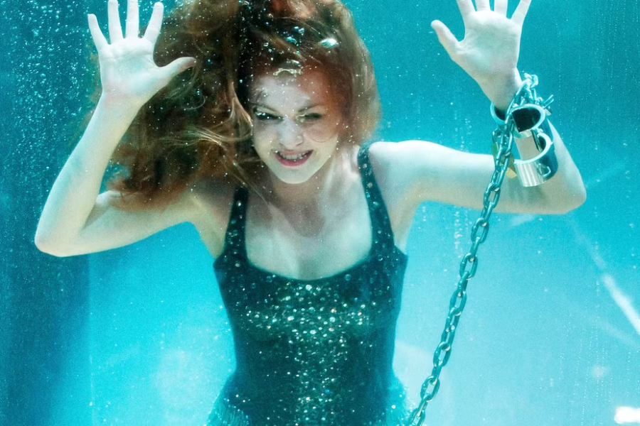 A red-haired woman in a sparkly black dress is submerged underwater with her hands against the glass and her wrist chained.