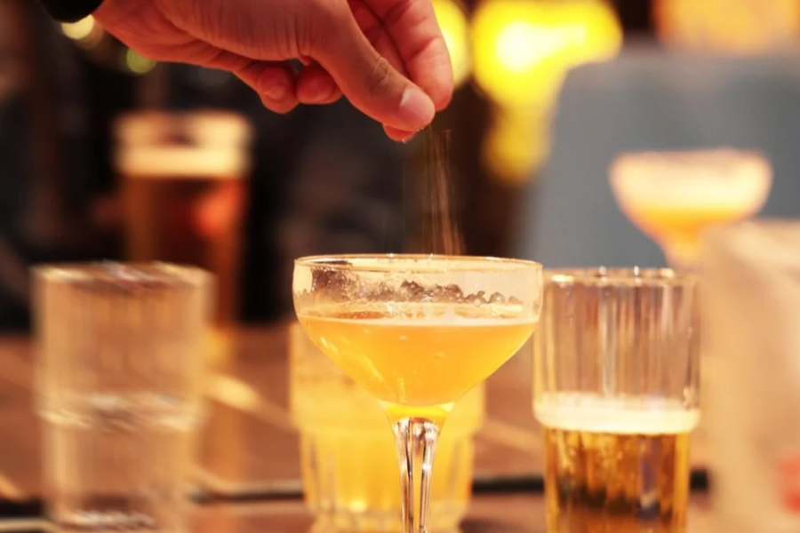 Close-up of a hand discreetly dropping powder into a yellow cocktail on a table surrounded by other drinks.