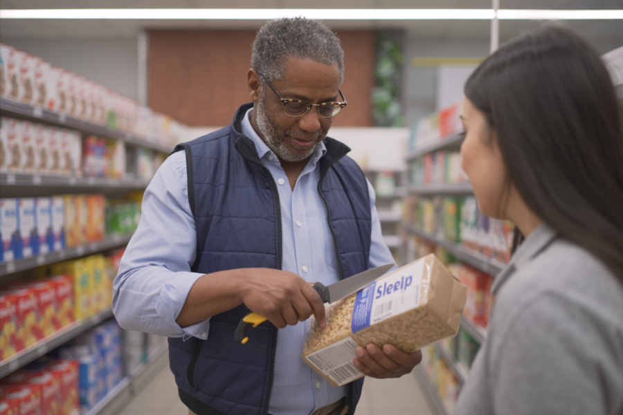 Man in a grocery store points to a package of pasta while speaking with a woman in the cereal aisle.