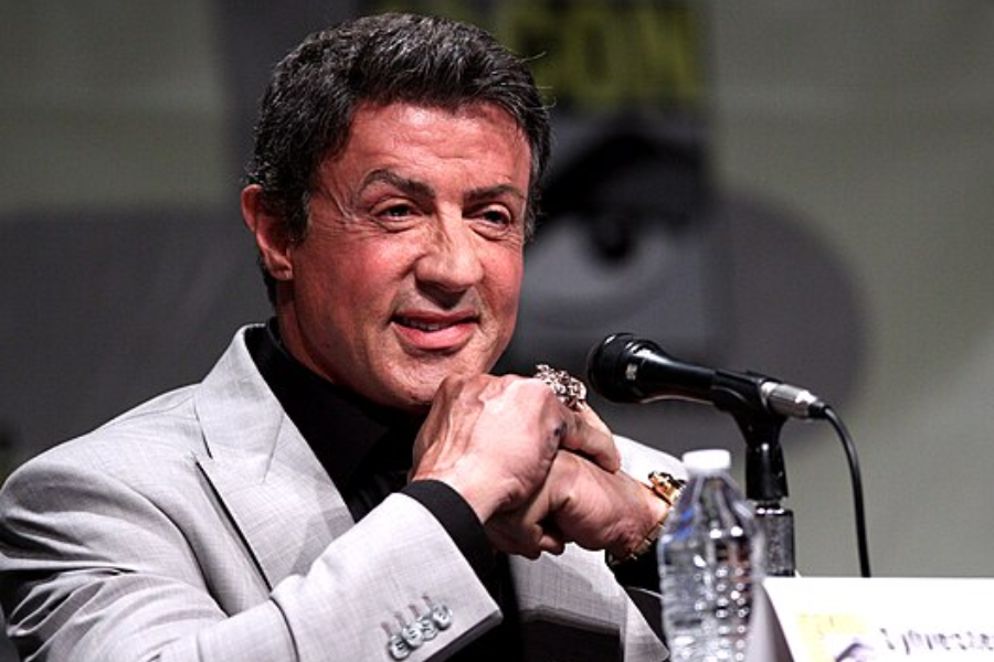 Man in a grey blazer speaks at a microphone during a panel event, smiling with his hands clasped.