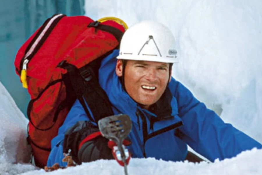 A mountaineer in a blue jacket and white helmet grips an ice tool while ascending through snow, carrying a large red backpack with climbing gear.