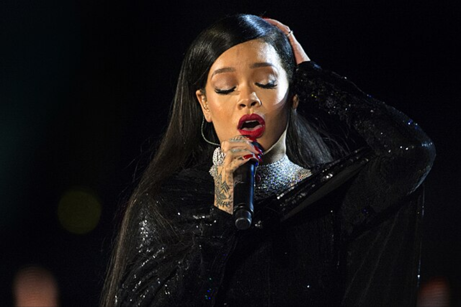 Woman with long dark hair and red lipstick sings into a microphone in a sequined black outfit, hand raised to her head.