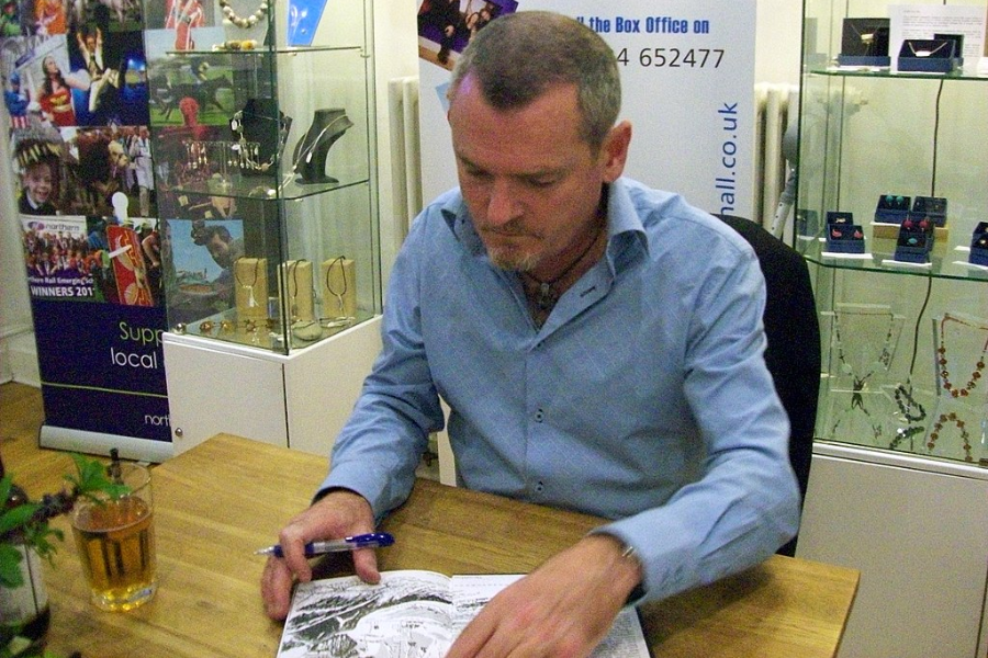 A man with a pen signs a book while seated at a wooden table in a shop, surrounded by glass cases of jewelry and display banners.