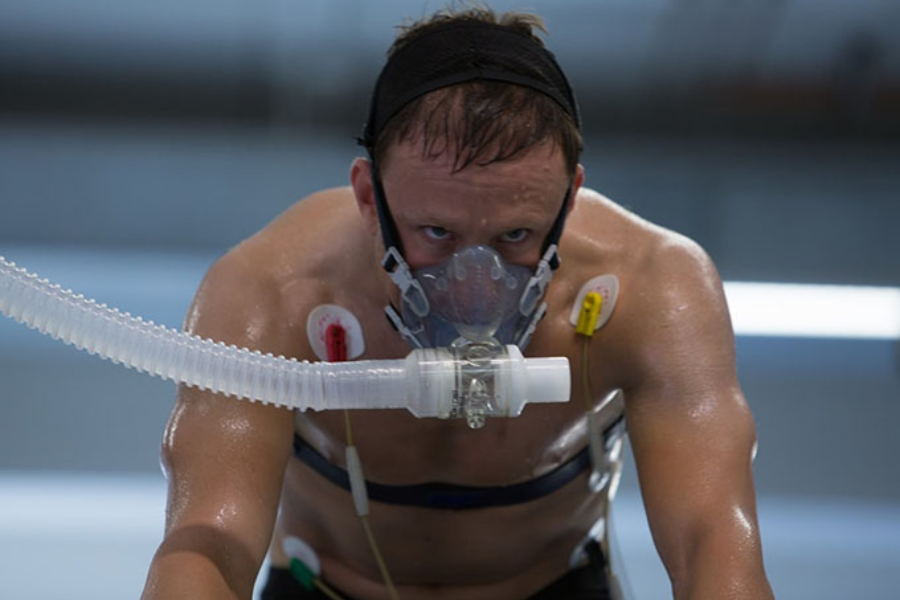 A shirtless man in a lab setting wears a breathing mask and chest sensors while intensely pedaling an exercise bike.