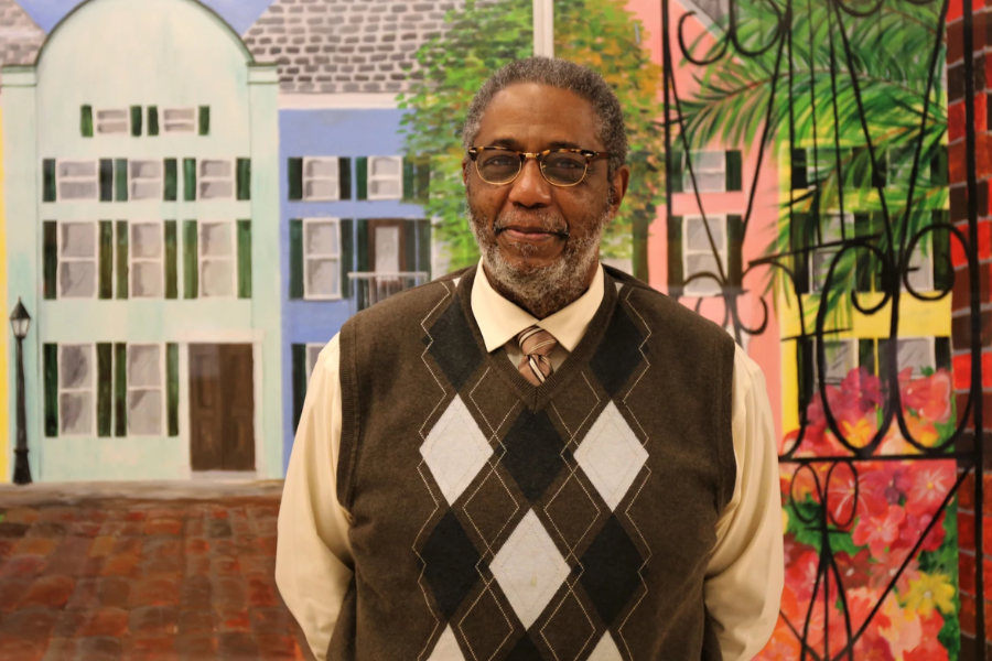 Henry Darby stands smiling in front of a colorful mural depicting pastel buildings and greenery.