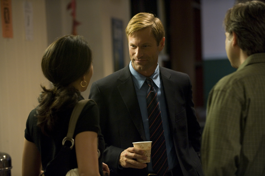 A man in a suit and tie holds a coffee cup while smiling and conversing with two other people in a dim hallway.