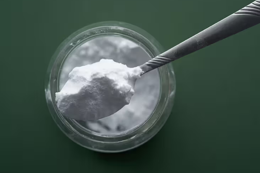 Overhead view of a spoonful of white baking soda above a clear glass jar on a dark green background.