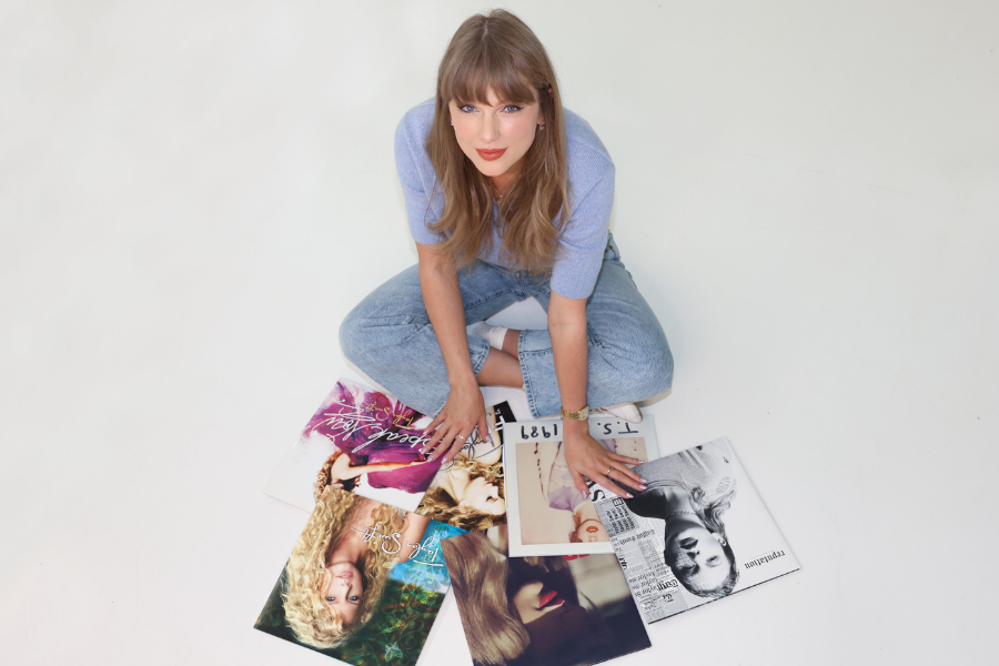 Woman in a lavender top and jeans sits cross-legged on the floor surrounded by multiple vinyl records featuring her own album covers.