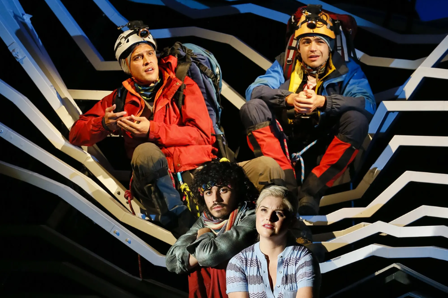A stage performance of Touching the Void shows two actors in mountaineering gear perched on a stylized mountain set, with two others seated below.