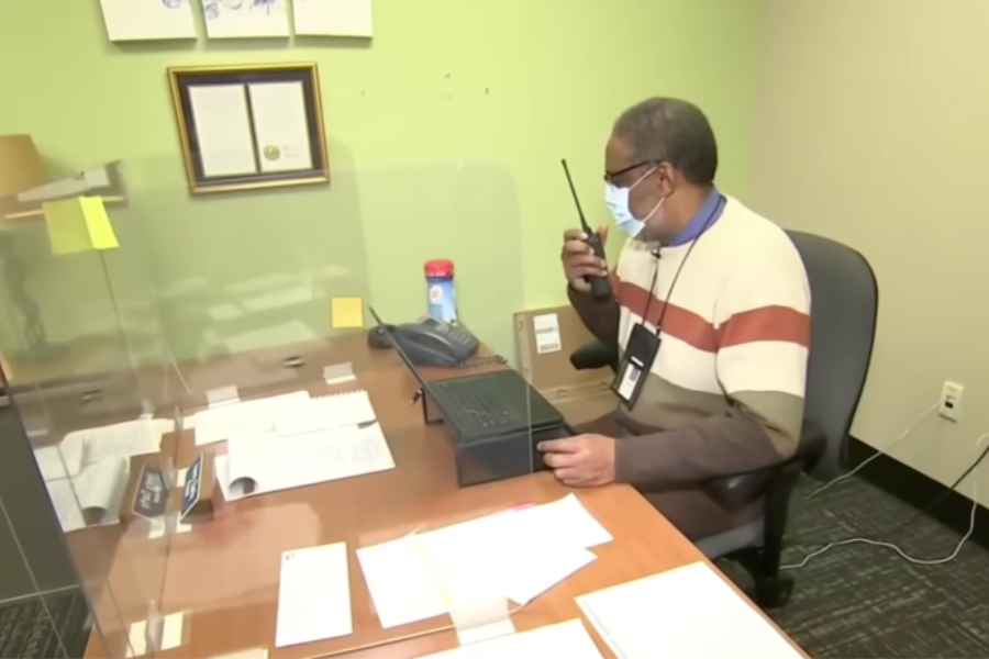 Henry Darby sits at a desk behind a clear plastic divider, speaking into a walkie-talkie while working.