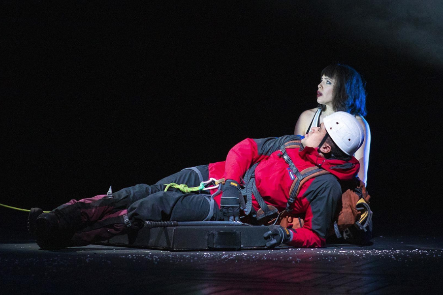 On stage, an actor in a red climbing jacket and helmet lies down with a woman in a dress sitting behind him, lit dramatically against a dark backdrop.