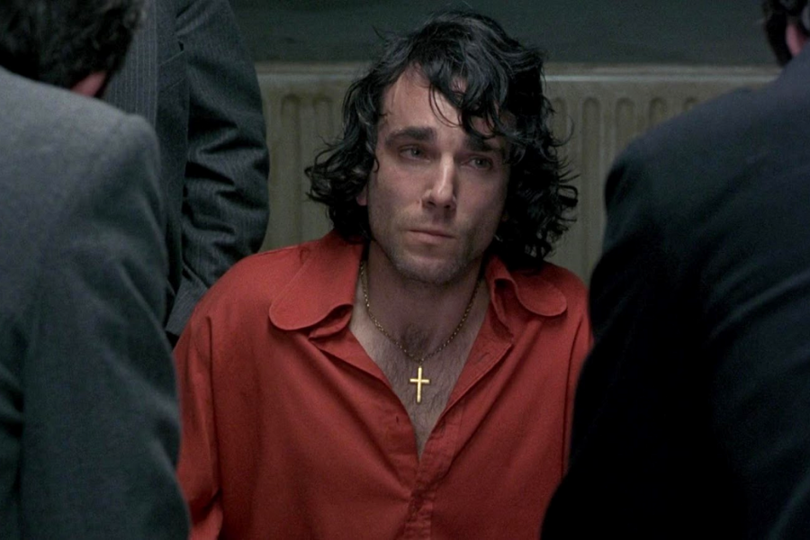 A man with long dark hair and a gold cross necklace sits in an interrogation room wearing a red shirt, looking tired and emotional.