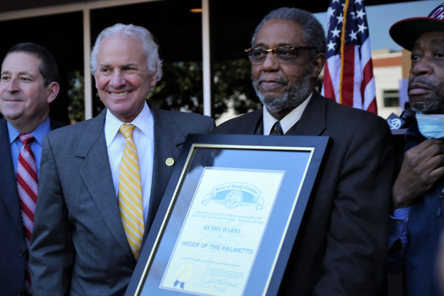Henry Darby holds a framed “Order of the Palmetto” award while standing next to state officials and smiling supporters.