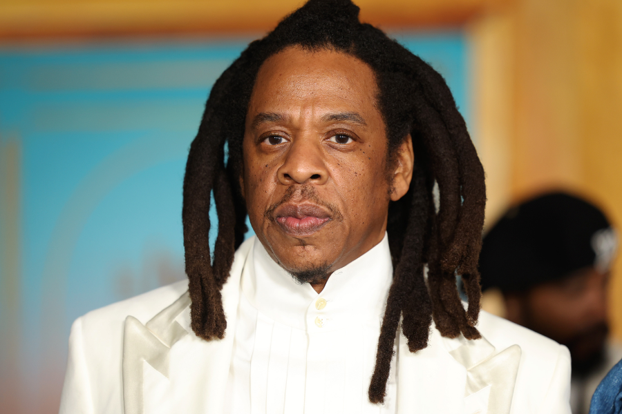Man in a formal white suit with long dreadlocks poses seriously in front of a soft-colored background.