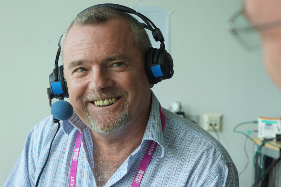 A man smiles while wearing a headset microphone in a studio or commentary box, with visible wires and a media lanyard around his neck.