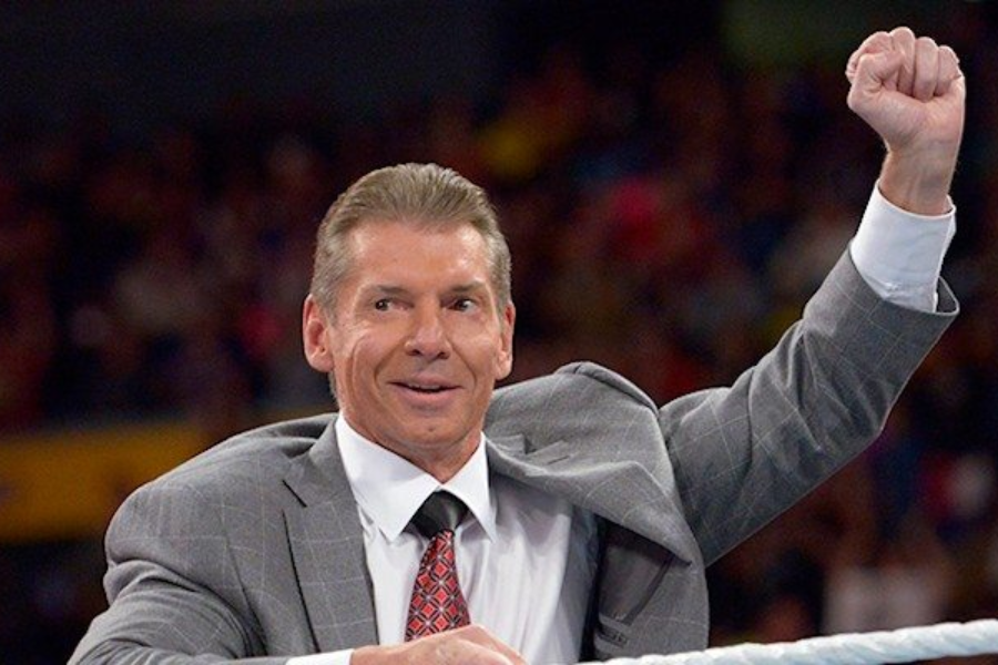 Man in a gray suit raises his fist while standing in a wrestling ring with a crowd in the background.