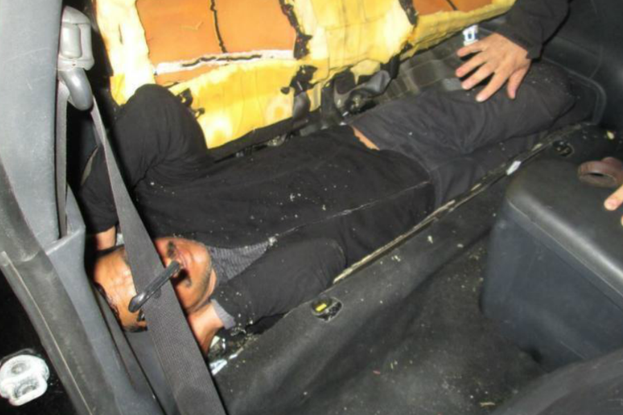 Man hidden behind a torn car seat back, lying in a tight space beneath it, revealing an improvised compartment.