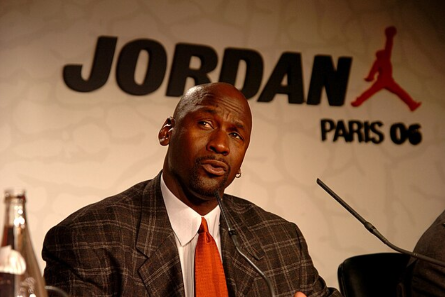 Man in a plaid suit and red tie sits at a press conference table with the word "Jordan" and "Paris 06" behind him.