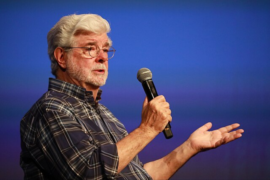Bearded man in glasses and a plaid shirt gestures with one hand while holding a microphone onstage.