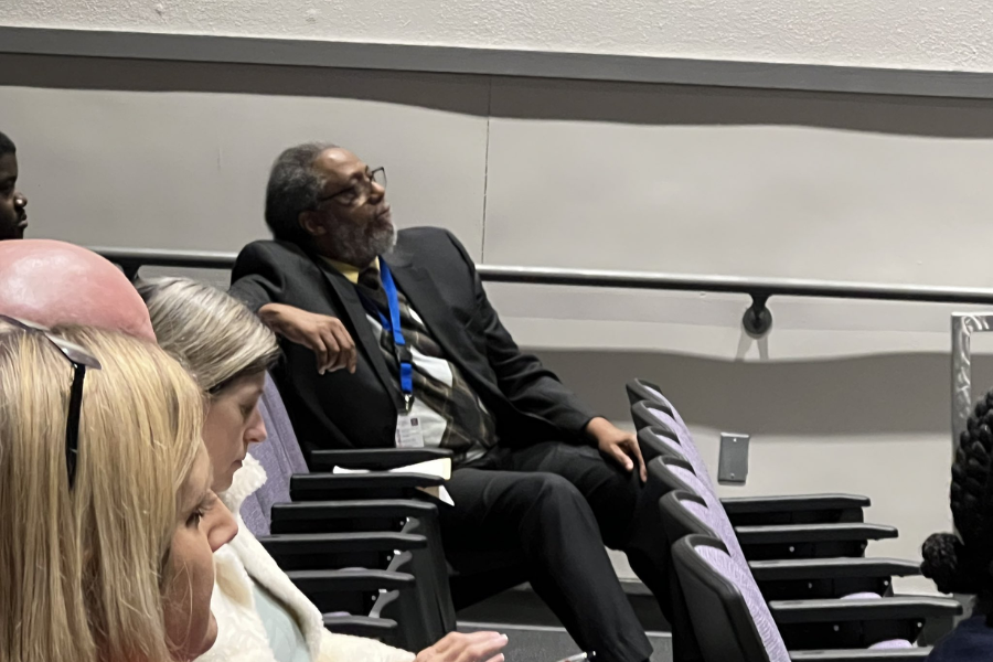 Man in a suit leans back in a chair while watching an event in an auditorium, surrounded by other seated attendees.